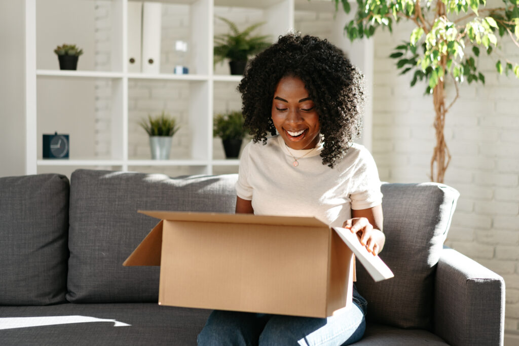 Smiling women on a couch opens a cardboard box from a delivery.